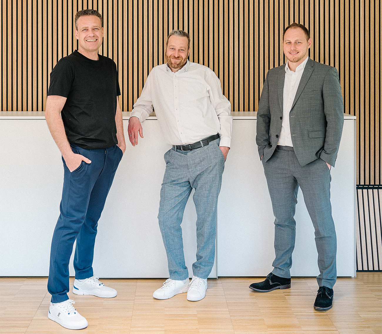 Die drei Geschäftsführer von Stiefvater & Walk – Magnus Walk, Björn Stiefvater und Steffen Erbsland – stehen entspannt nebeneinander vor einer Wand mit Holzlamellen